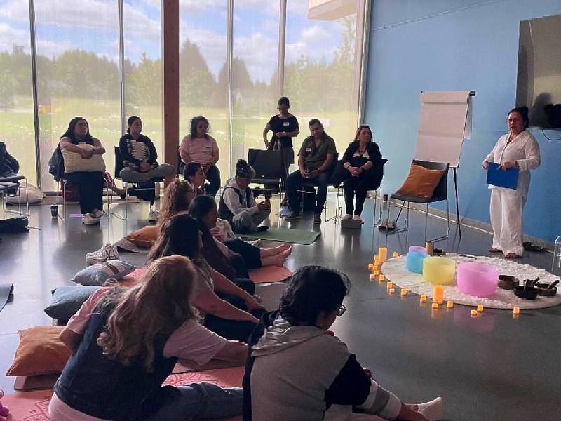 A group meeting in a lovely indoor space, several participants are seated in chairs, with pillows, others are on mats on the floor