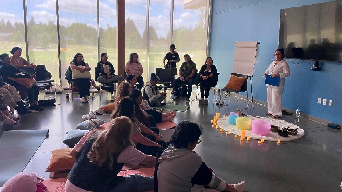 A group meeting in a lovely indoor space, several participants are seated in chairs, with pillows, others are on mats on the floor